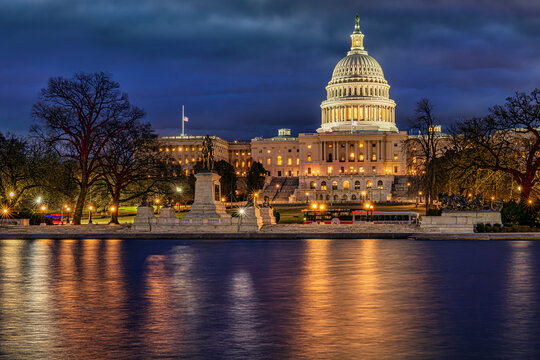 Illuminated US Capitol building at dusk reflecting on the water, framed by trees. Washington DC, WA, USA
