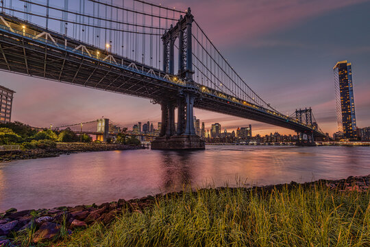 Cityscape with a lit suspension bridge over calm water at sunset, tall buildings in the background. New York City, NY, USA Williamsburg Bridge over East River