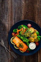 Fried salmon steak with boiled broccoli and mini carrots, fried potatoes and mayonnaise on wooden table. Top view