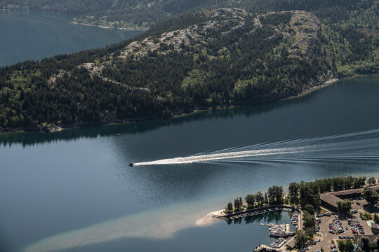 Aerial view of a lake with a speedboat creating a long trail near a forested mountain. Waterton Park, Alberta, Canada