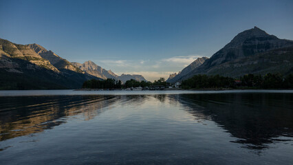 A serene mountain lake at sunset with clear skies and lush greenery reflecting on the water. Waterton Park, Alberta, Canada