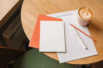Close-up overhead view of a blank paper card, to do list, notebook, pen and pencil, cup of cappuccino on a table.