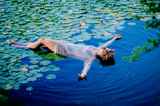 Woman in a white dress and flower crown floats peacefully on a lily pad-covered pond. Wildcat Lake Park, WA, USA