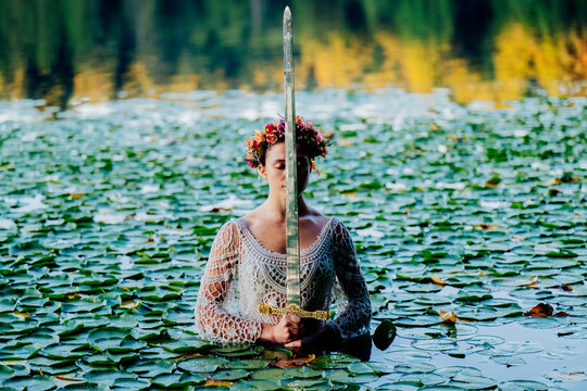 Woman in lace dress and flower crown holds a sword in a lily-covered lake. Wildcat Lake Park, WA, USA