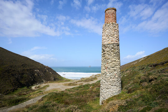 Old tin mine and Trevellas Porth beach, near Blue Hills, St Agnes, Cornwall, UK. Tall stone chimney amidst coastal landscape with hills and ocean under a blue sky.