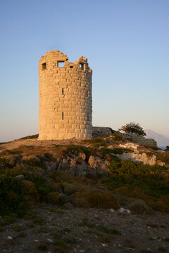The ancient Greek tower of Drakonon, Ikaria island, Greece. Ancient stone watchtower on a hill at sunset with clear sky and rugged landscape.