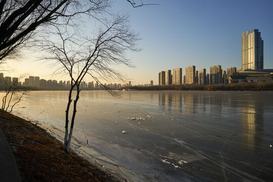 Frozen river with distant city skyline and bare trees at sunset.