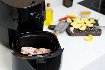 Air fryer with raw chicken wings, potato wedges and spices on white marble countertop, closeup