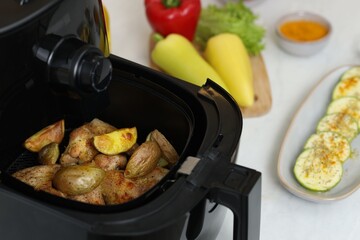 Air fryer with grilled chicken wings, potato wedges, vegetables and spices on white marble table, closeup