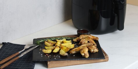 Air fryer with grilled chicken wings, potato wedges and spices on white marble countertop, closeup