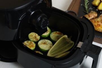 Air fryer with grilled vegetables, chicken wings, potato wedges and spices on white table, closeup