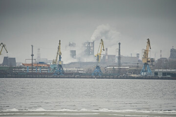 Industrial landscape, a factory with pipes and cranes in the port against a gray foggy sky