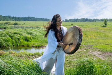 Beautiful female shaman in a trance in the white dress drumming in the natural environment