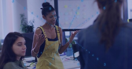 Woman in yellow overalls guiding team around desk in office, with monitors and sticky note screen