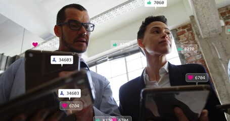 Colleagues in suits holding tablets and viewing social media stats in office with floating icons