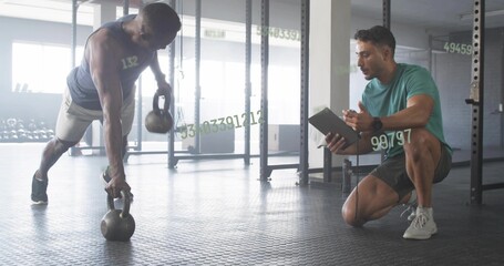 Performing renegade row man holding two kettlebells at gym with trainer tablet, copy space