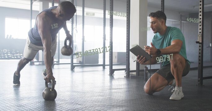 Performing renegade row man holding two kettlebells at gym with trainer tablet, copy space