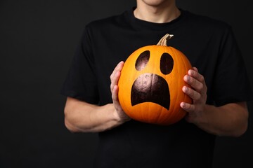 Halloween celebration. Man holding pumpkin with drawn spooky face on black background, closeup