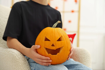 Halloween celebration. Boy holding pumpkin with carved spooky face in armchair indoors, closeup