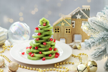 Christmas tree made of kiwi, pomegranate seeds and festive decor on beige table against light background with blurred lights, closeup. Bokeh effect