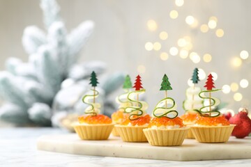Christmas food. Tasty tartlets with red caviar, cream cheese, cucumber and decor on white marble table against background with blurred lights, closeup. Bokeh effect
