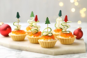 Christmas food. Tasty tartlets with red caviar, cream cheese, cucumber and decor on white marble table against background with blurred lights, closeup. Bokeh effect
