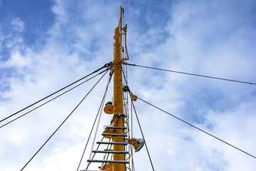 A bright yellow ship mast framed against a clear blue sky, with multiple rigging lines, a metal ladder, and various hardware projecting from the mast. Nautical engineering and industrial design.