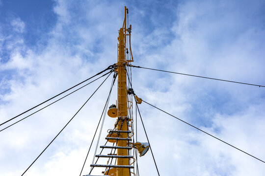 A bright yellow ship mast framed against a clear blue sky, with multiple rigging lines, a metal ladder, and various hardware projecting from the mast. Nautical engineering and industrial design.