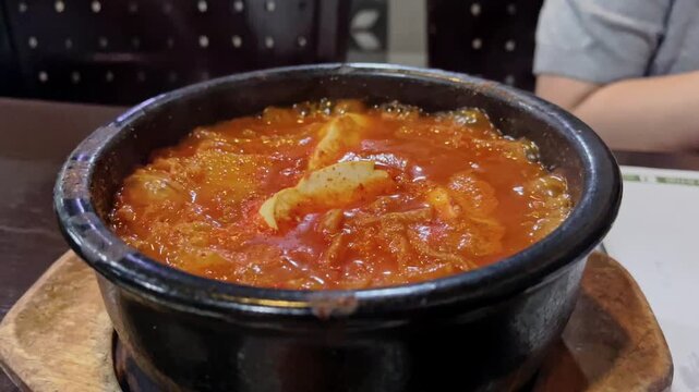 Traditional Korean kimchi jjigae bubbling in hot stone bowl, with blurred woman chatting in restaurant background