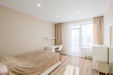 Cozy bedroom featuring a bed, desk, and natural light from a large window. Neutral color palette with modern lighting and minimalist decor