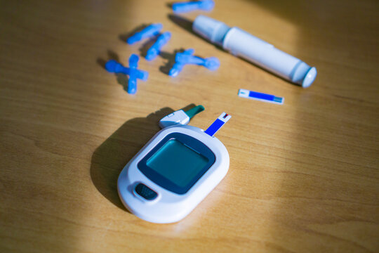 small white glucometer device with a blue test strip inserted lying on a wooden table, lancet pen and several blue single use lancets scattered nearby, concept of healthcare, medical technology
