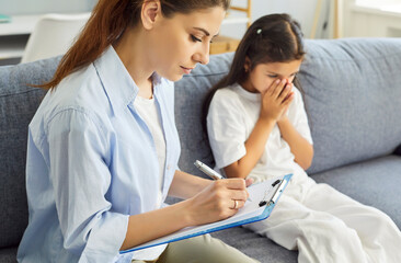 Psychologist notes in child therapy session. Beside her, the child cries into hands, anxiety rising...