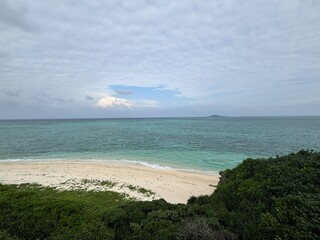 A stunning view of the emerald green "Miyako Blue" ocean and a pristine white sand beach on Miyako Island, Okinawa, Japan, with lush green vegetation in the foreground under a cloudy sky
