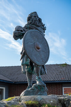 King Harald, Fairhair statue, bronze statue of a bearded man holding a shield stands on a rocky pedestal in front of a brick building under a clear blue sky, Haugesund, Norway