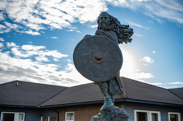 King Harald, Fairhair statue, bearded warrior holding a shield stands in front of a house under a partly cloudy blue sky, Haugesund, Norway