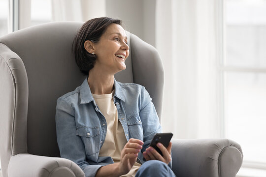 Happy dreamy mature female use smartphone relaxing in cozy armchair - Powered by Adobe