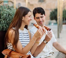 Happy couple  eating a pastry croissant, smiling and looking away on a city street