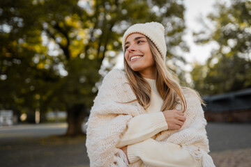 attractive smiling young blond woman walking in winter park having fun in warm white knitted sweater, cape and hat, cold season