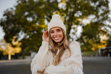 attractive smiling young blond woman walking in winter park having fun in warm white knitted sweater, cape and hat, cold season