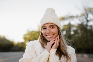 attractive smiling young blond woman walking in winter park having fun in warm white knitted sweater, cape and hat, cold season