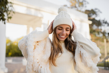 attractive smiling young blond woman walking in winter park having fun in warm white knitted sweater, cape and hat, cold season