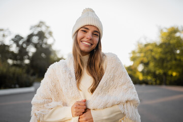 attractive smiling young blond woman walking in winter park having fun in warm white knitted sweater, cape and hat, cold season