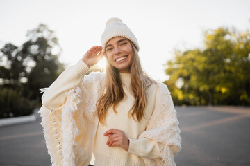 attractive smiling young blond woman walking in winter park having fun in warm white knitted sweater, cape and hat, cold season