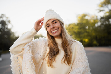 attractive smiling young blond woman walking in winter park having fun in warm white knitted sweater, cape and hat, cold season