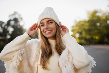 attractive smiling young blond woman walking in winter park having fun in warm white knitted sweater, cape and hat, cold season