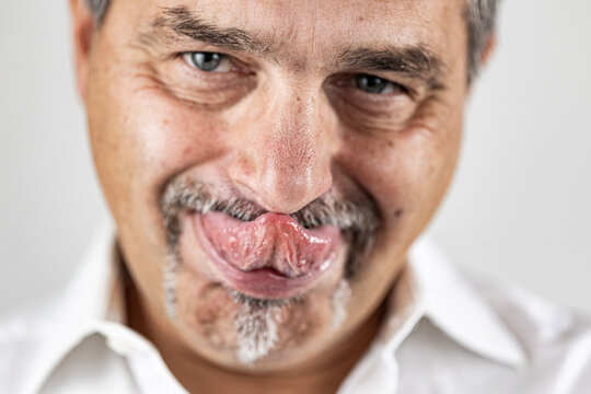 Mature man making a humorous face while touching his nose with his tongue. Captured in bright, natural light, highlighting skin texture, gray facial hair, and a playful mood..