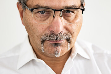 Close-up image of a confident mature man wearing eyeglasses and a white shirt, featuring detailed facial hair and a calm, assertive expression.
