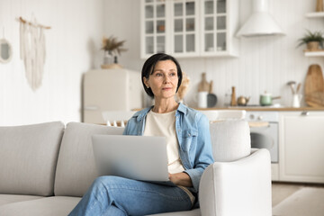 Pensive older lady sitting on couch with notebook looking away