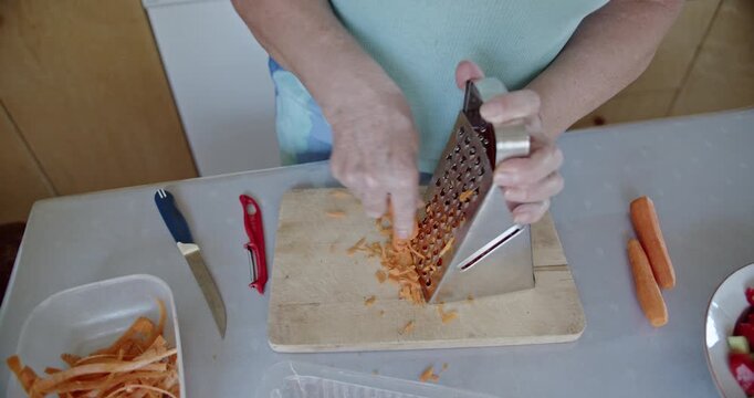 Close-up of a person's hands grating fresh, vibrant orange carrots with a metal grater on a wooden cutting board. The scene highlights healthy food preparation and home cooking in a bright kitchen.