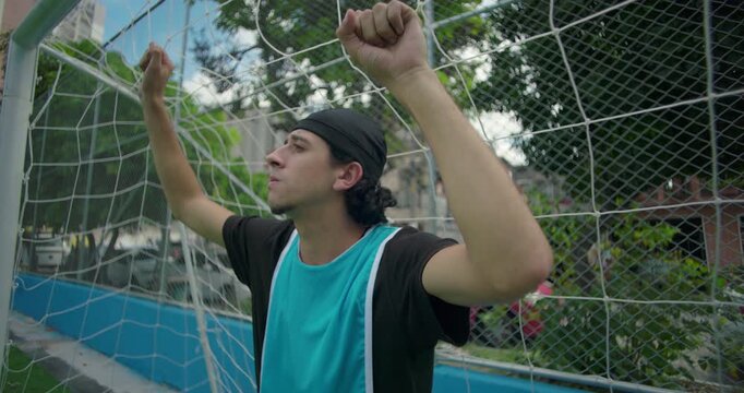 Hispanic soccer player stands pensively against goal net on quiet urban field, lost in thought after game, reflecting on life, purpose, and personal moments of solitude